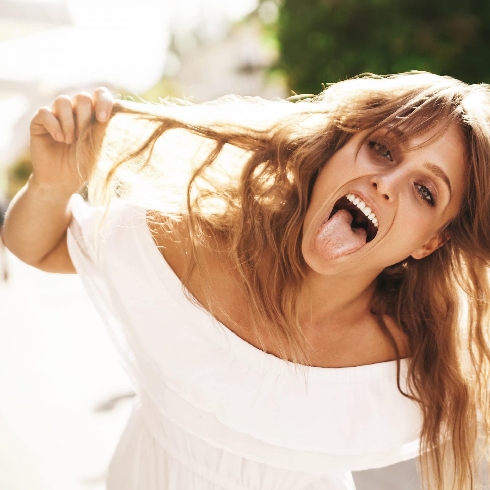 Portrait of cheerful blonde hipster girl without makeup going crazy making funny face and showing her tongue on the street background. Touching her hair